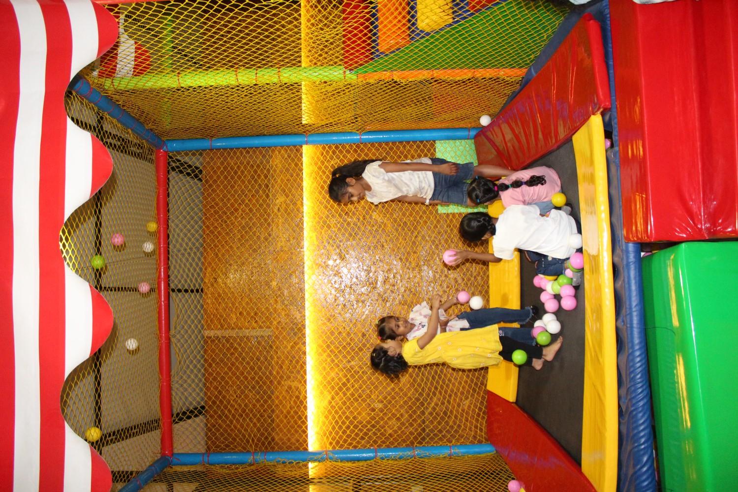 Children playing inside a colorful indoor trampoline enclosure with safety netting and plastic balls