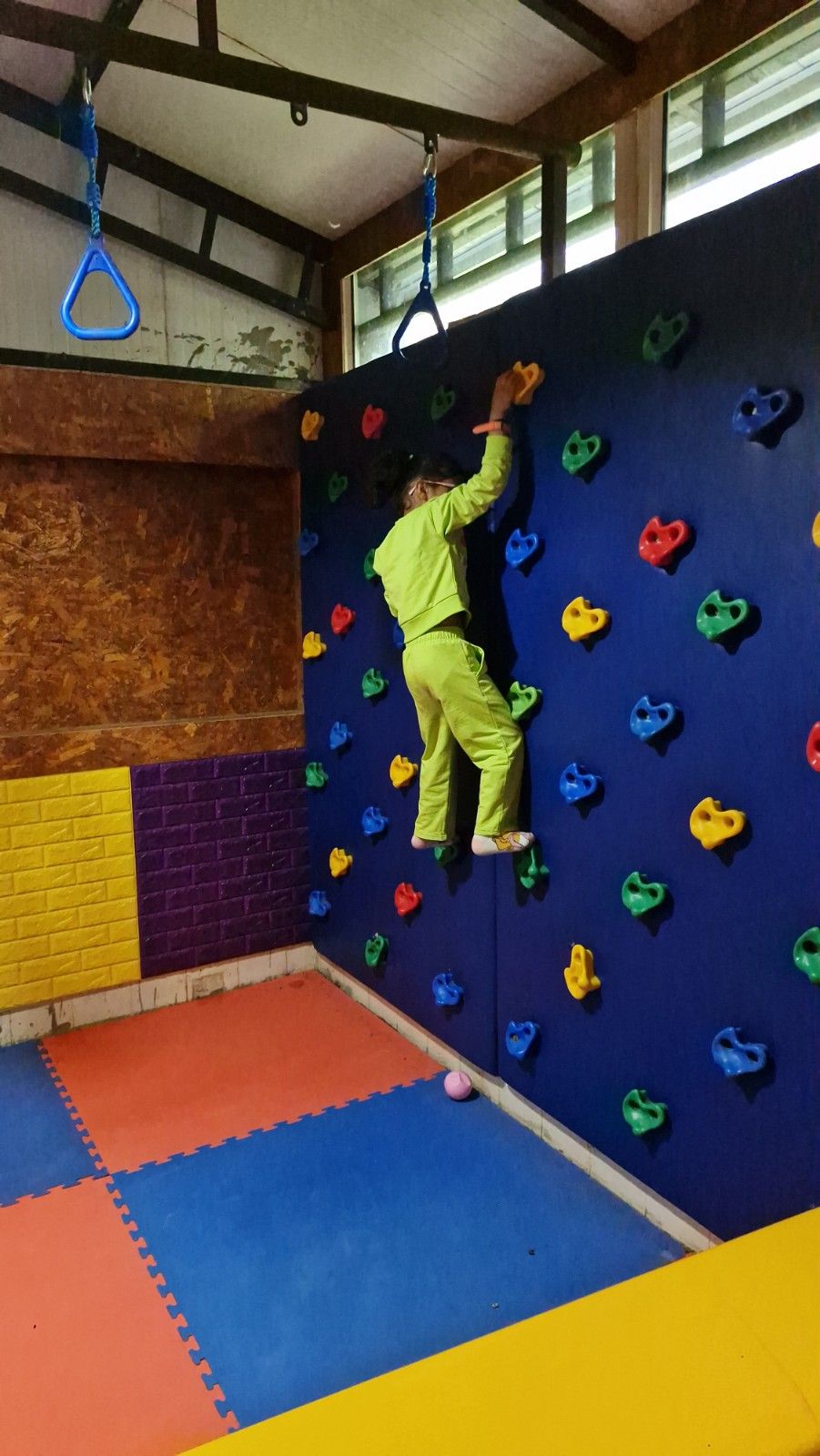 A young girl riding a green plastic rocker toy in an indoor play area with padded walls