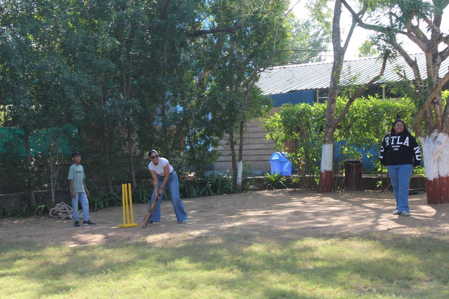 A group of people playing a game of cricket in a park setting surrounded by trees.