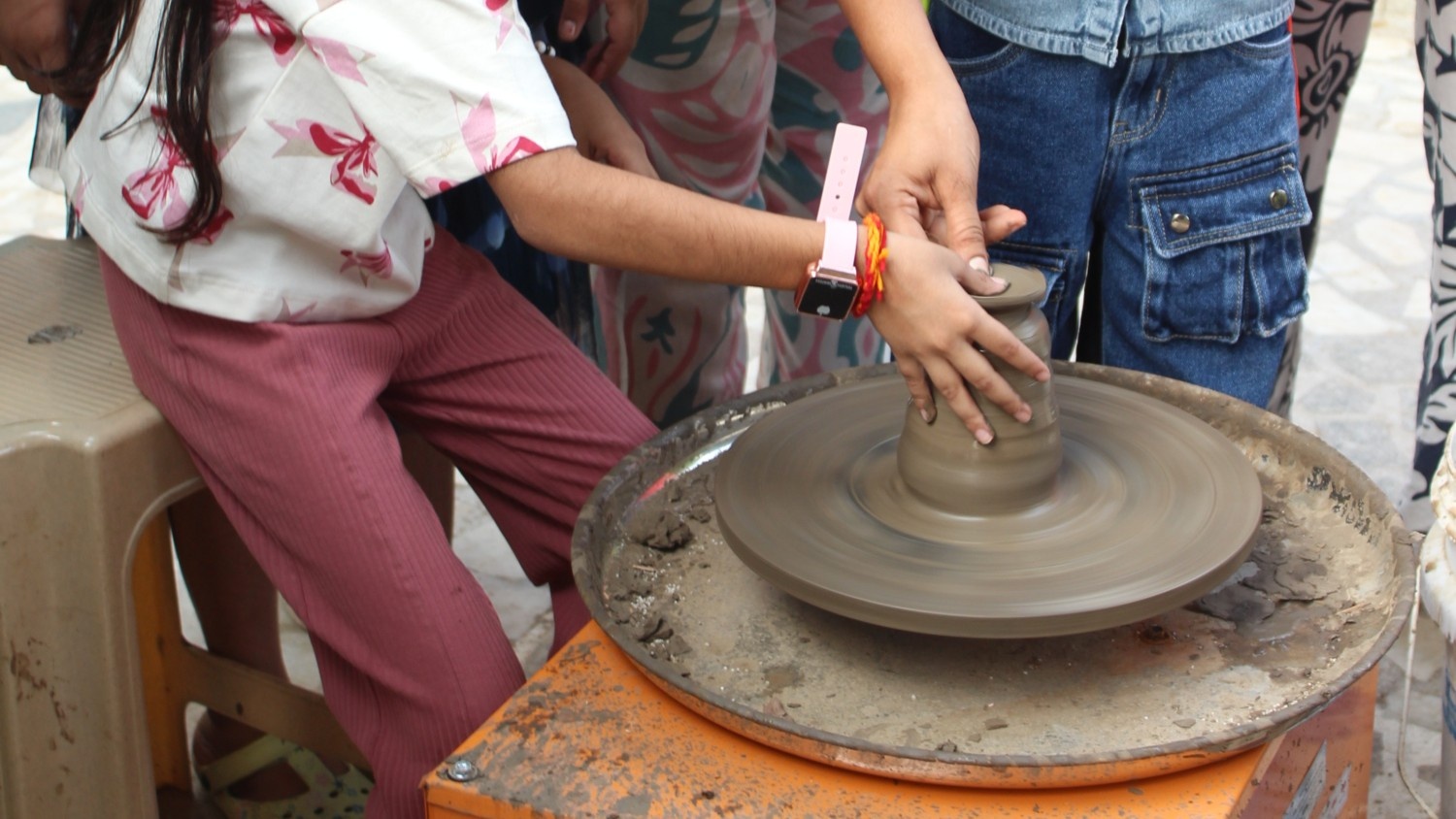 A close-up of a child's hands being guided by an instructor to shape clay on a spinning pottery wheel.