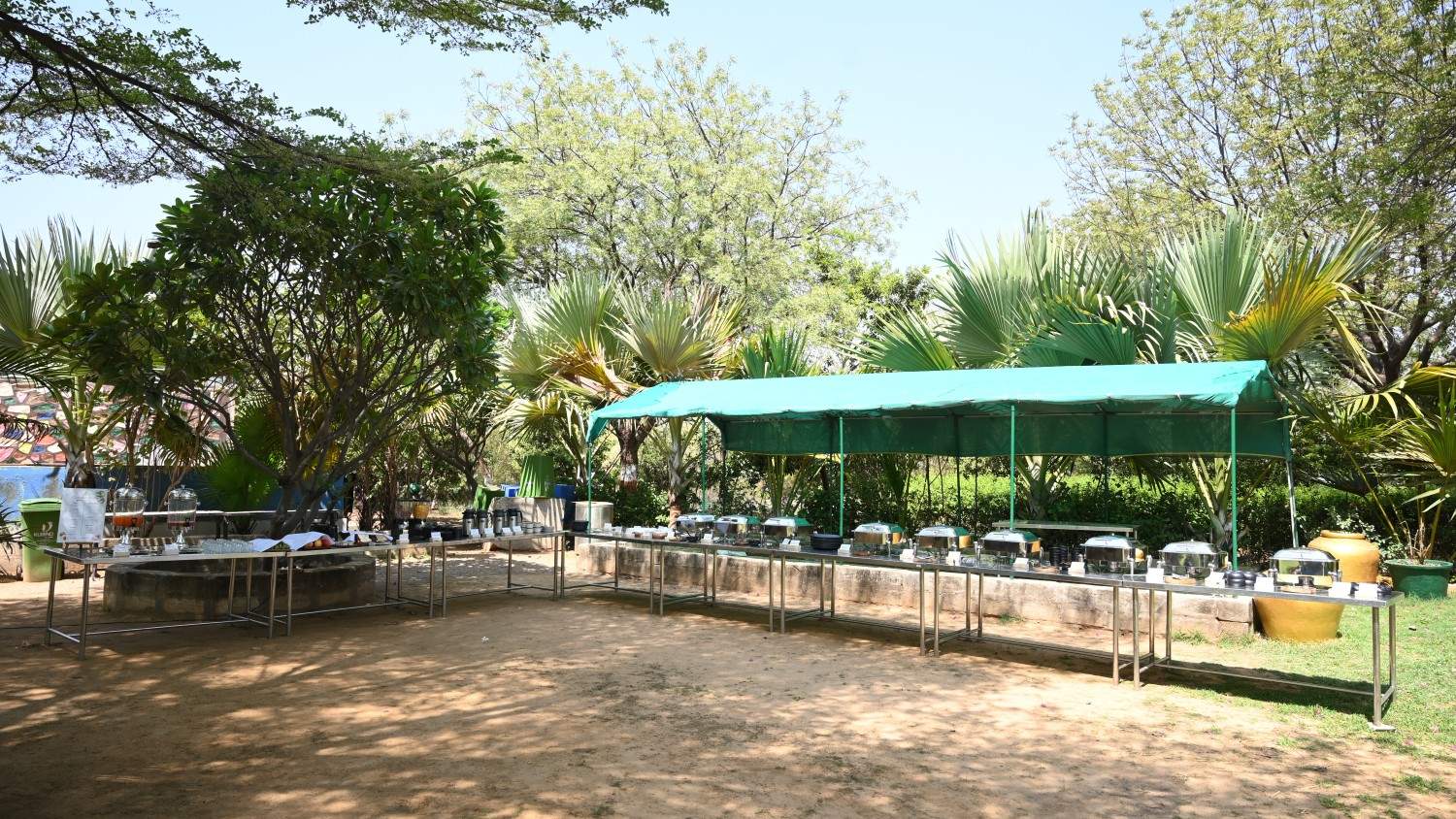 Outdoor buffet setup with food counters under a green canopy surrounded by trees and greenery.