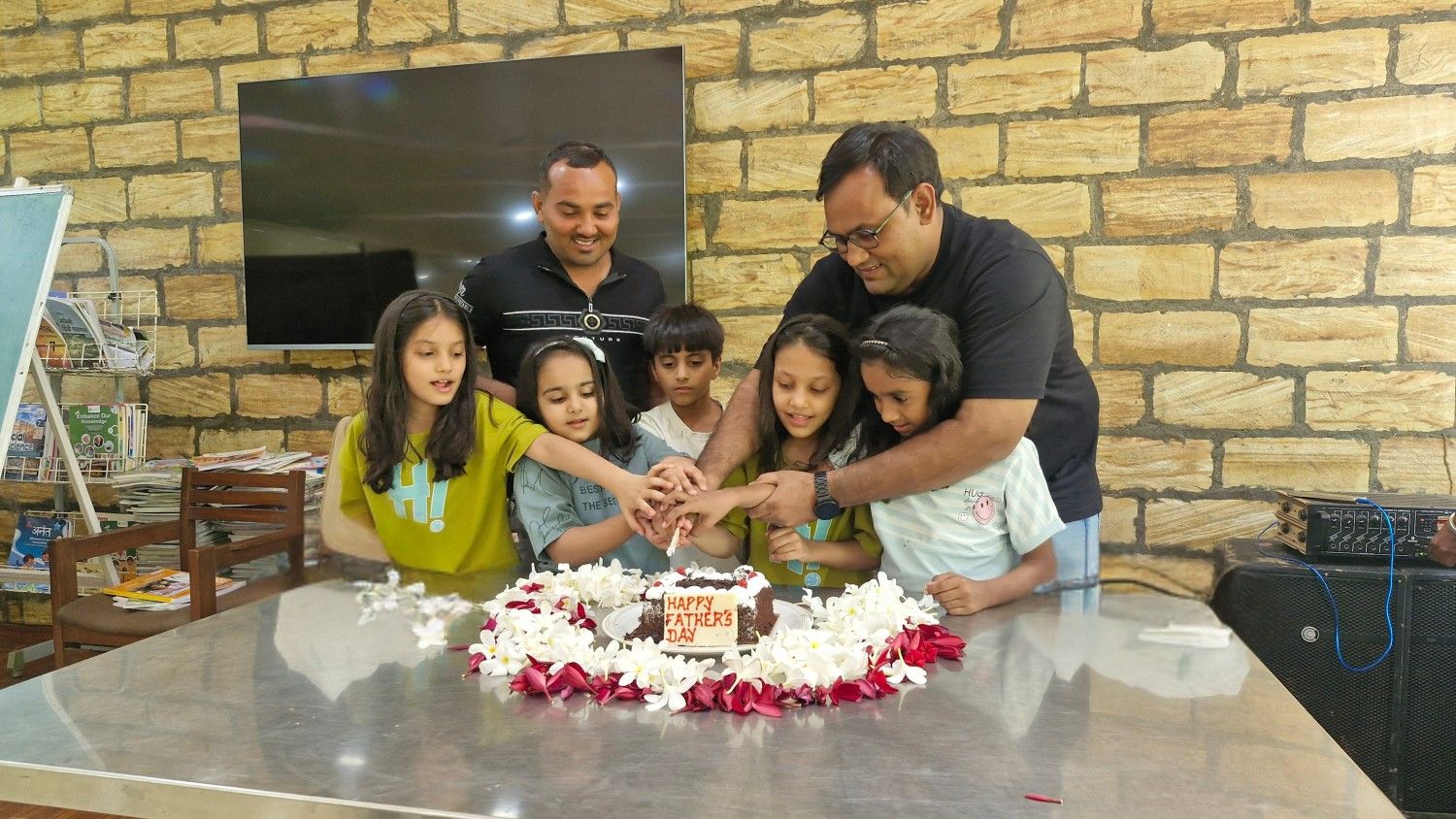 Group of children and two adults cutting a cake that says Happy Father’s Day on a decorated table.
