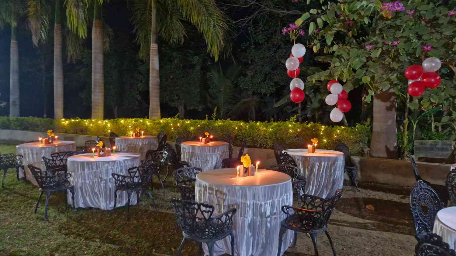 Outdoor restaurant seating area at night featuring round tables with white linens, candles, and string lights in the background