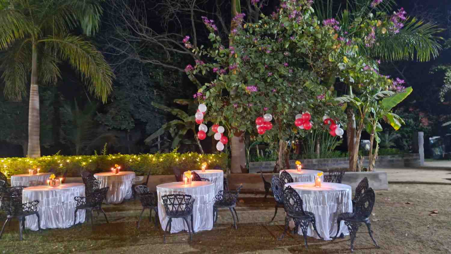 Romantic outdoor dinner setup with round tables, white tablecloths, and red and white balloons hanging from trees